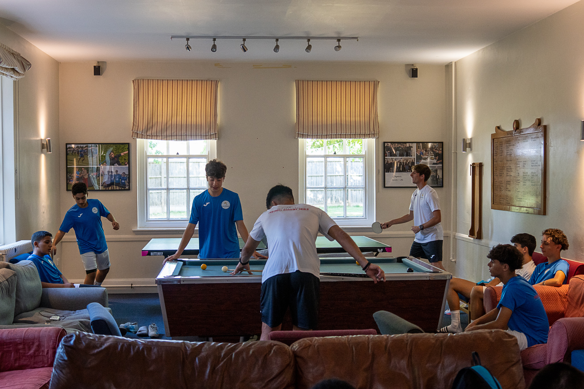 Sala común de la residencia, con sofás de cuero y butacas alrededor y fotografías enmarcadas en las paredes. En primer plano, dos jugadores adolescentes juegan al billar sobre una mesa de madera oscura con tapete azul claro y cantos metálicos: uno, de espaldas, se inclina sobre la mesa con el taco de madera en la mano, con la bola blanca y una amarilla sobre el tapete. Justo detrás hay una mesa de ping-pong verde distinta, con el logotipo de Butterfly en el canto, y un tercer jugador con camiseta blanca sosteniendo una pala. Otros chicos miran sentados.