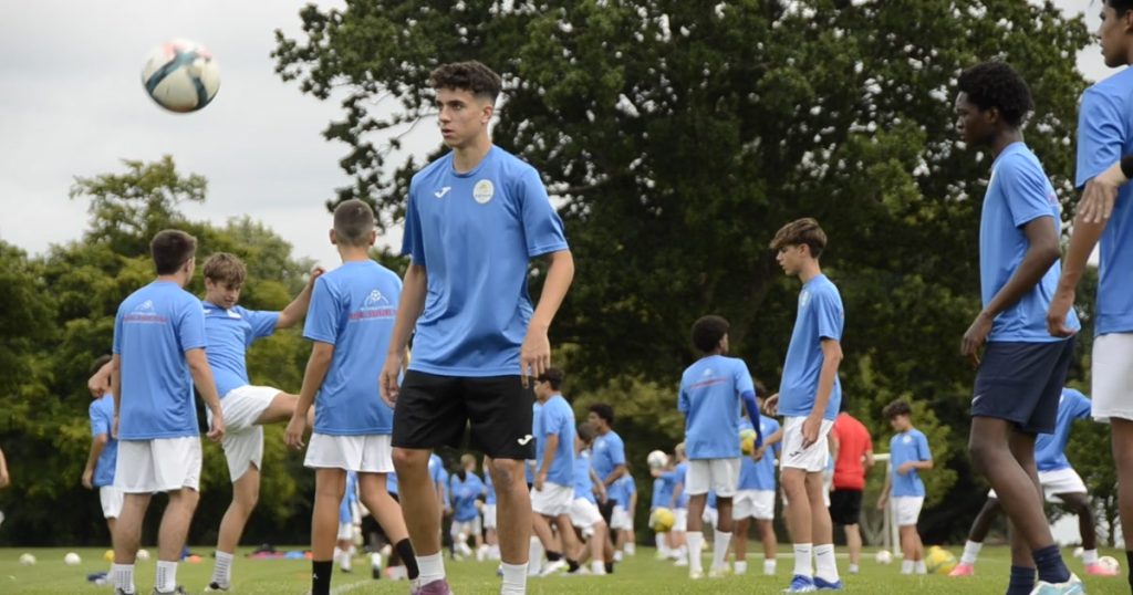 Un grupo de jugadores jovenes entrena con balon sobre hierba en un campamento de futbol de Ertheo; en primer plano uno controla el balon y detras decenas de companeros trabajan en la misma sesion.