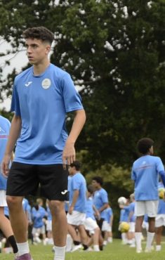 Un grupo de jugadores jovenes entrena con balon sobre hierba en un campamento de futbol de Ertheo; en primer plano uno controla el balon y detras decenas de companeros trabajan en la misma sesion.